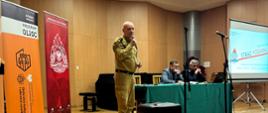 Funkcjonariusz Państwowej Straży Pożarnej w umundurowaniu w kolorze piaskowym prowadzi wystąpienie na scenie, trzymając mikrofon. Za nim przy stole siedzą dwaj mężczyźni przedstawiciele miasta Sosnowiec oraz Policji. Na scenie znajdują się roll-upy dotyczące programu OLiOC – Ochrona Ludności i Obrona Cywilna oraz Państwowej Straży Pożarnej, a także ekran z prezentacją. Wystąpienie odbywa się w ramach szkolenia poświęconego przygotowaniu kadry oświatowej do reagowania na zagrożenia.