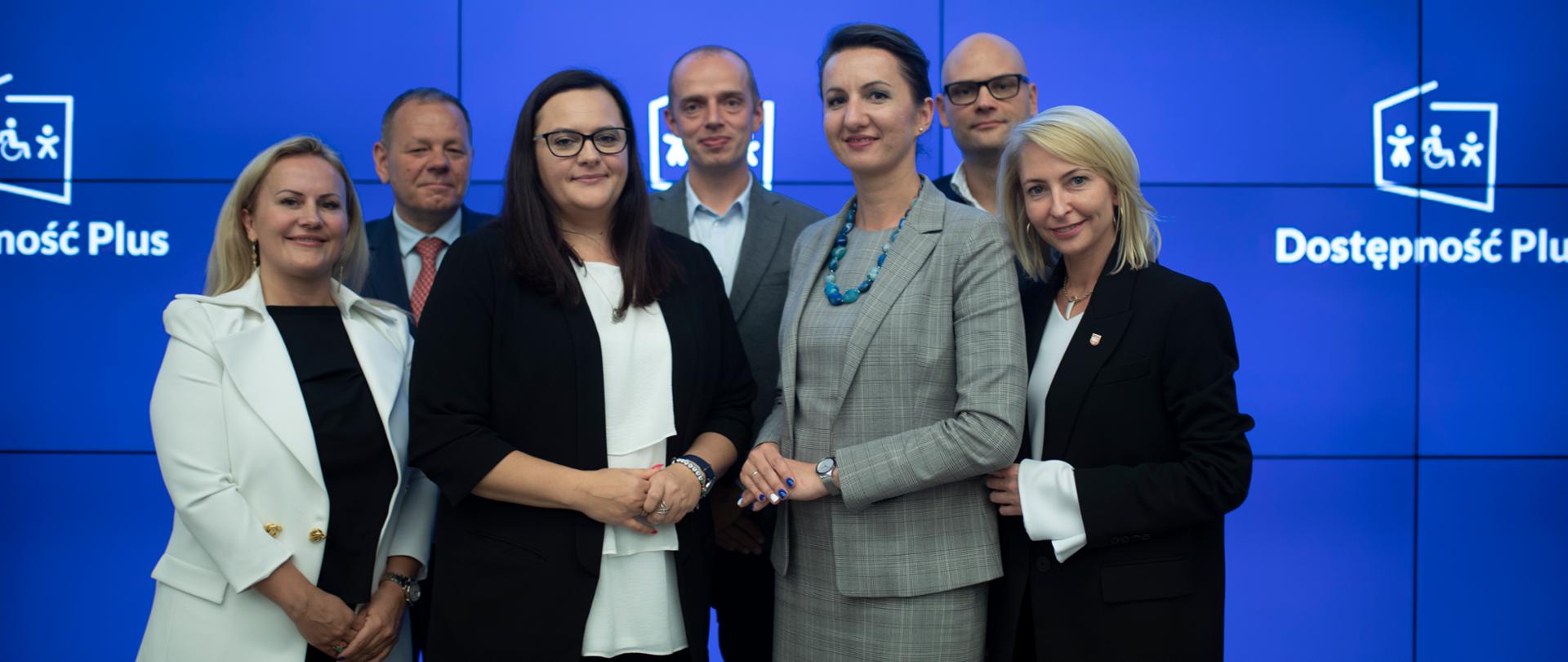 Siedem osób na scenie, z przodu cztery, z tyłu trzy, z przodu druga od lewej stoi wiceminister Jarosińska-Jedynek, z tyłu niebieskie tło z logo programu Dostępność Plus