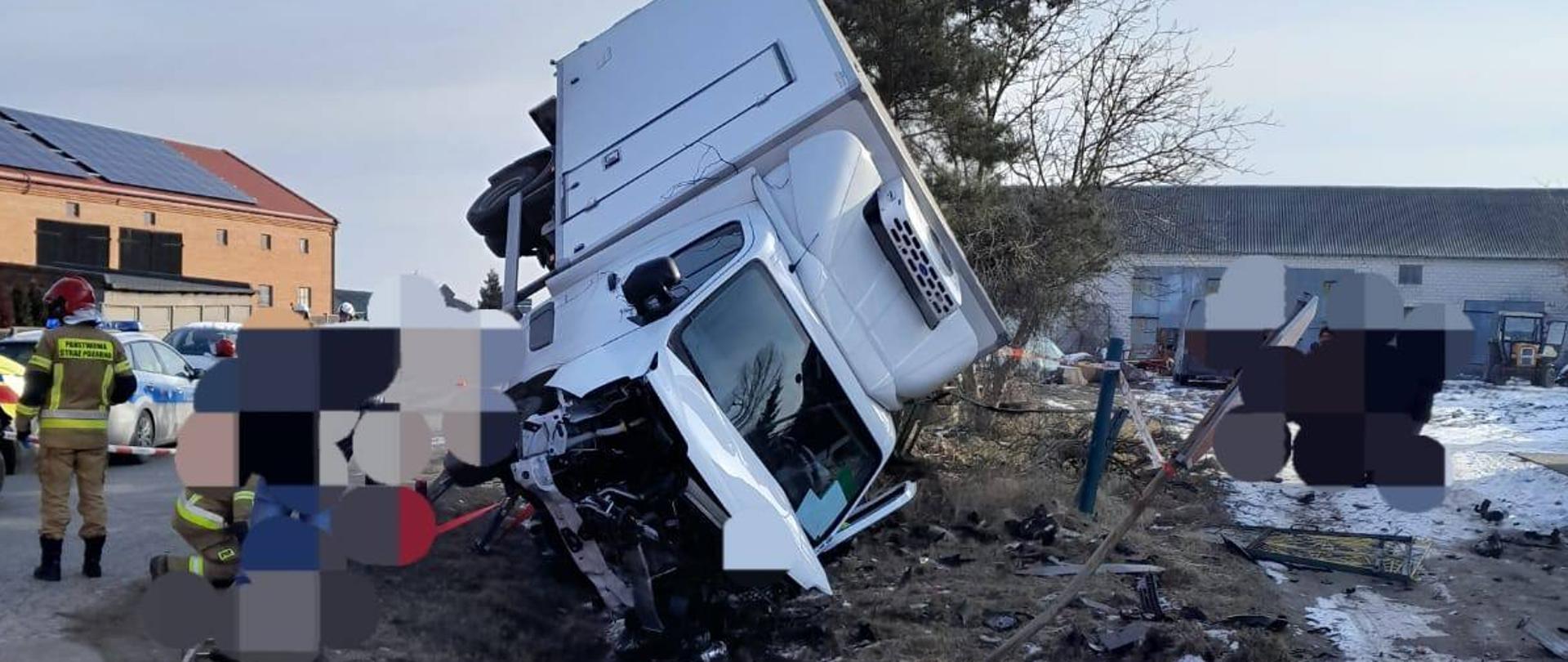 Na zdjęciu widać biały samochód dostawczy, którego przód jest w rowie a tył wisi w powietrzu. Obok stoi strażak w żółtym stroju i czerwonym hełmie. Za strażakiem widać samochód policji. Dookoła porozrzucane są elementy karoserii oraz uszkodzonego ogrodzenia. W tle widać budynki gospodarcze. Jest widno.