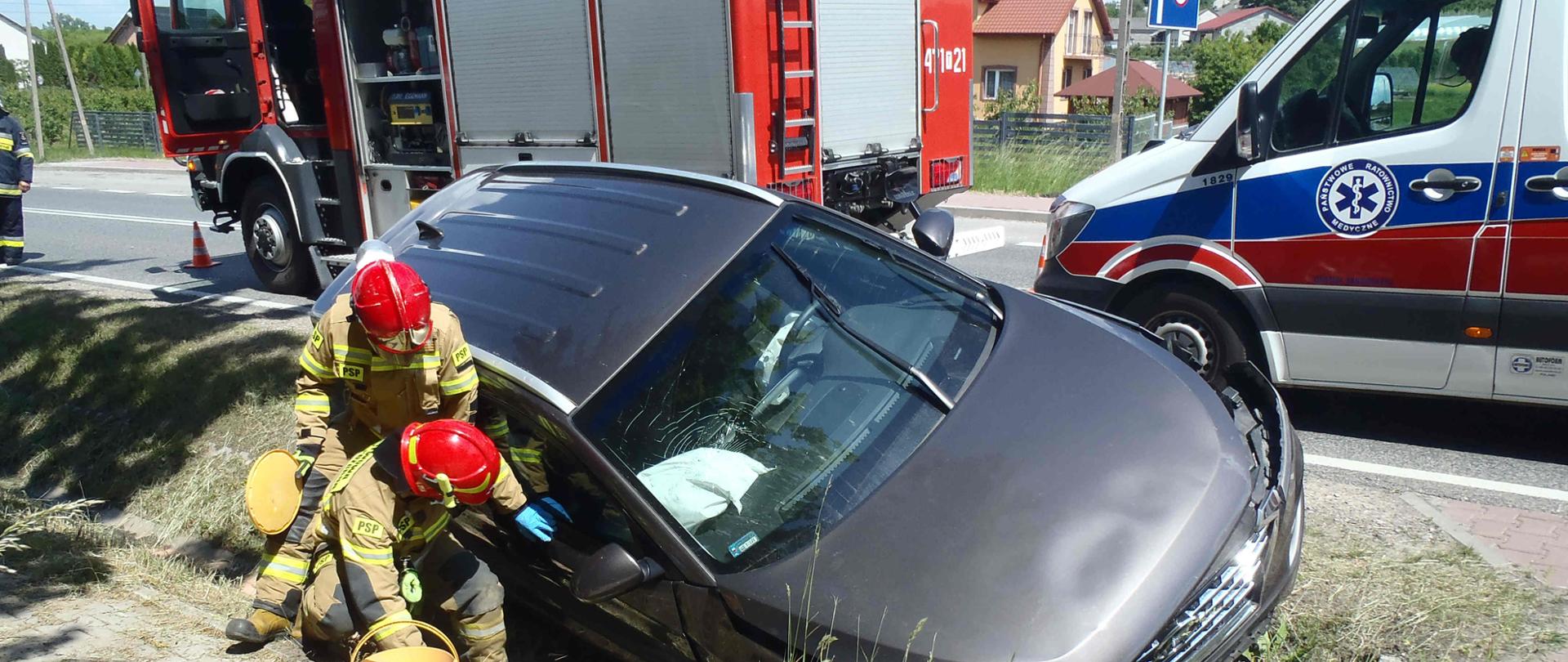 Zdjęcie zrobione na tle pojazdów ratowniczych. Na zdjęciu widać rozbity samochód przy którym stoi strażak. W oddali widać samochód strażacki. Na drodze ustawiony stożek ostrzegawczy.