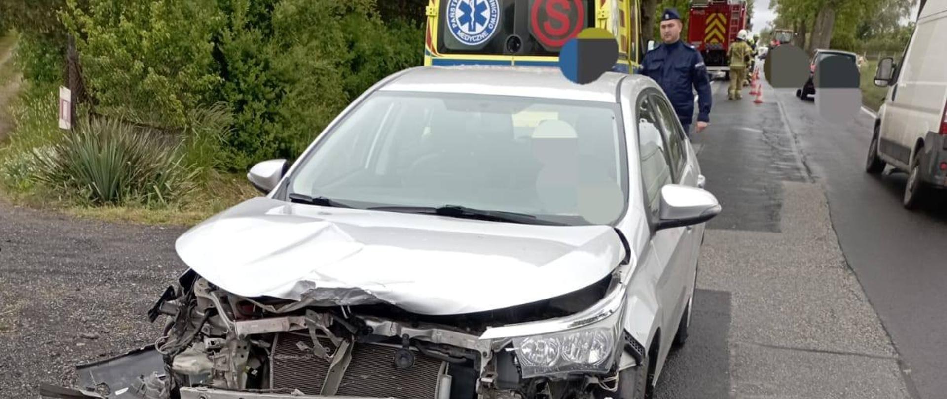 Na zdjęciu widać rozbity srebrny samochód osobowy, który ma uszkodzony przód m.in. pogiętą maskę, oderwany zderzak. Pojazd stoi na poboczu asfaltowej drogi, częściowo na pasie jezdni. Za samochodem stoi żółta karetka pogotowia ratunkowego obok której idzie policjant. Za karetką widoczny jest samochód straży pożarnej oraz strażacy ubrani w stroje w kolorze piaskowym i białe hełmy. W tle widać krzewy i drzewa. W prawym górnym rogu widoczne są inne pojazdy przejeżdżające nobok miejsca wypadku. Jest widno.
