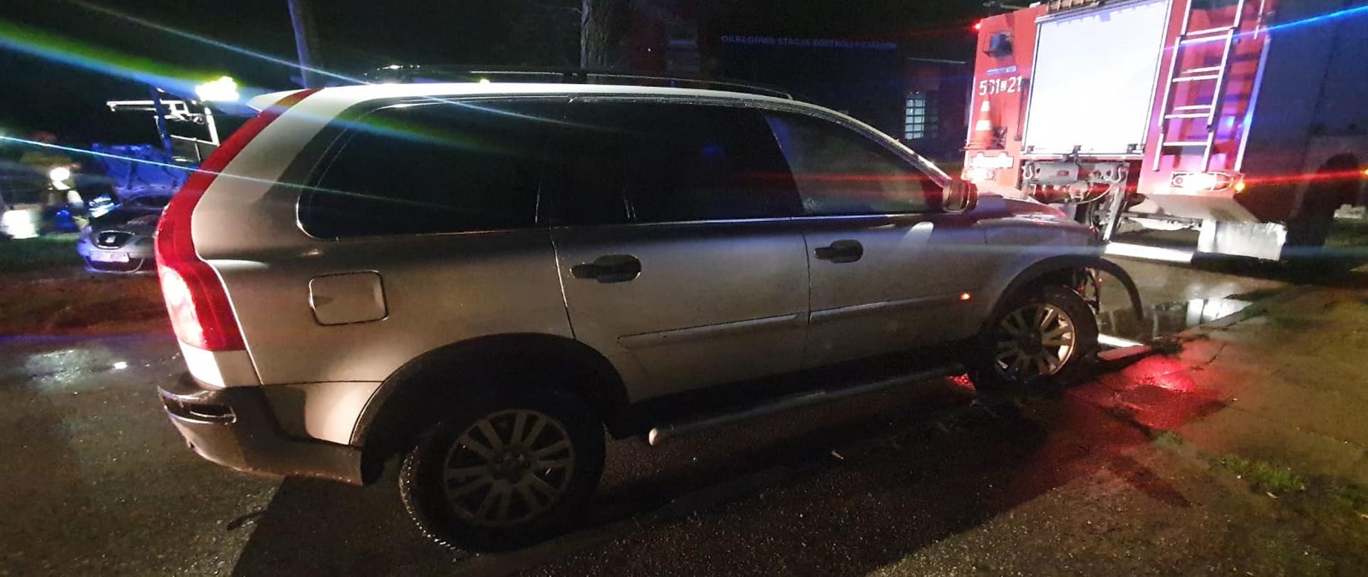 Zdjęcie przedstawia w porze wieczornej samochód koloru srebrnego. Ma uszkodzony przód pojazdu z prawej strony. Przed pojazdem stoi straż pożarna.