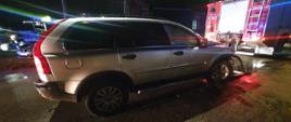 Zdjęcie przedstawia w porze wieczornej samochód koloru srebrnego. Ma uszkodzony przód pojazdu z prawej strony. Przed pojazdem stoi straż pożarna.