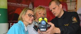 Targi edukacyjne. Sala sportowa w ZSZ w Rawiczu. Strażak prezentuje 2 kobietom kamerę termowizyjną. W tle ścianka reklamowa.