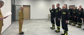 Trzech strażaków z uniesioną prawą ręką składa ślubowanie Komendantowi Miejskiemu. Za nimi w szeregu stoją strażacy z podziału bojowego w ubraniu koszarowym. 