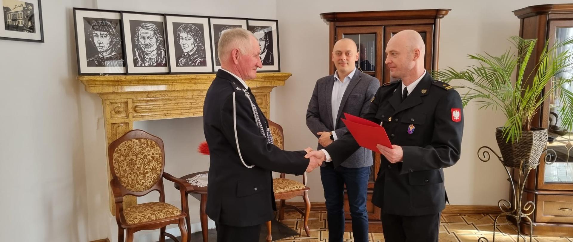 Na zdjęciu widać komendanta powiatowego Państwowej Straży Pożarnej w Jędrzejowie mł. bryg. Mariusza Stolarczyka, który wręcza druhowi Marianowi Zynek decyzję o przyznaniu świadczenia ratowniczego z tytułu wysługi lat w Ochotniczej Straży Pożarnej. Strażacy w umundurowaniu wyjściowym. Za strażakami stoi wójt gminy Słupia Pan Tomasz Koper. Zdjęcie z robione w Urzędzie Gminy Słupia Jędrzejowska.
