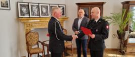 Na zdjęciu widać komendanta powiatowego Państwowej Straży Pożarnej w Jędrzejowie mł. bryg. Mariusza Stolarczyka, który wręcza druhowi Marianowi Zynek decyzję o przyznaniu świadczenia ratowniczego z tytułu wysługi lat w Ochotniczej Straży Pożarnej. Strażacy w umundurowaniu wyjściowym. Za strażakami stoi wójt gminy Słupia Pan Tomasz Koper. Zdjęcie z robione w Urzędzie Gminy Słupia Jędrzejowska.