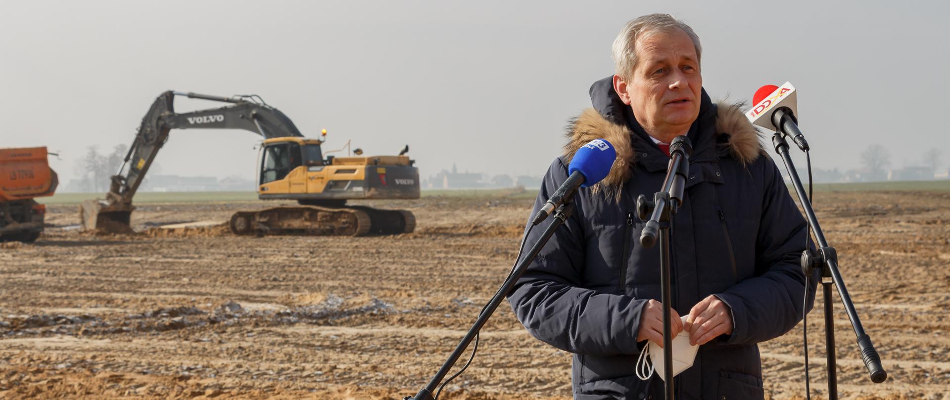 Przemówienie wojewody opolskiego Sławomira Kłosowskiego podczas uroczystego wkopania pierwszej łopaty pod budowę obwodnicy Olesna. Na zdjęciu stojący w prawej stronie kadru wojewoda, w grubej kurtce. Przed nim trzy mikrofony na statywie. Wojewoda spogląda na dziennikarzy. W tle budowa obwodnicy, po lewej stronie kadru koparka. 