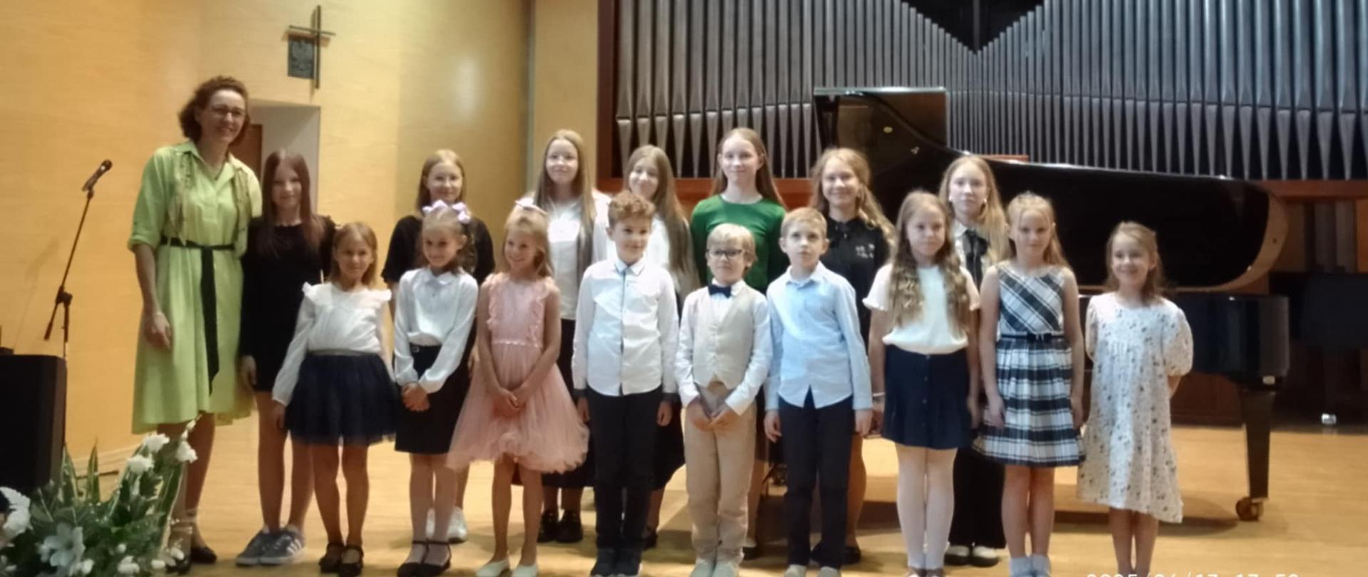 Zdjęcie przedstawia grupę dzieci i jedną dorosłą osobę, prawdopodobnie nauczycielkę, stojących na scenie w sali koncertowej.
W lewej części zdjęcia stoi uśmiechnięta kobieta w średnim wieku, ubrana w jasnooliwkową sukienkę z czarnym paskiem, z kręconymi włosami. Po jej prawej stronie stoi szereg dzieci w różnym wieku, od kilku do kilkunastu lat. Większość z nich jest elegancko ubrana. Dziewczynki mają na sobie sukienki lub spódniczki z bluzkami, a chłopcy koszule, niektórzy z kamizelkami. Dzieci są uśmiechnięte i patrzą w stronę obiektywu. Tło sceny jest drewniane, w kolorze jasnego brązu. Po prawej stronie widoczny jest duży, czarny fortepian koncertowy oraz fragment piszczałek organowych, tworzących ciemny, geometryczny wzór na ścianie. Na lewej ścianie, za plecami kobiety, wisi ciemny krzyż.
W lewym dolnym rogu zdjęcia widać fragment jasnej kompozycji kwiatowej, prawdopodobnie bukietu. 