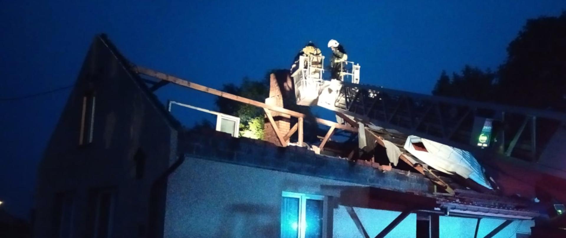 na zdjęciu widoczny jest parterowy budynek mieszkalny z zerwanym częściowo dachem. Dwóch strażaków w ubraniach specjalnych, z kosza drabiny zabezpiecza uszkodzony dach