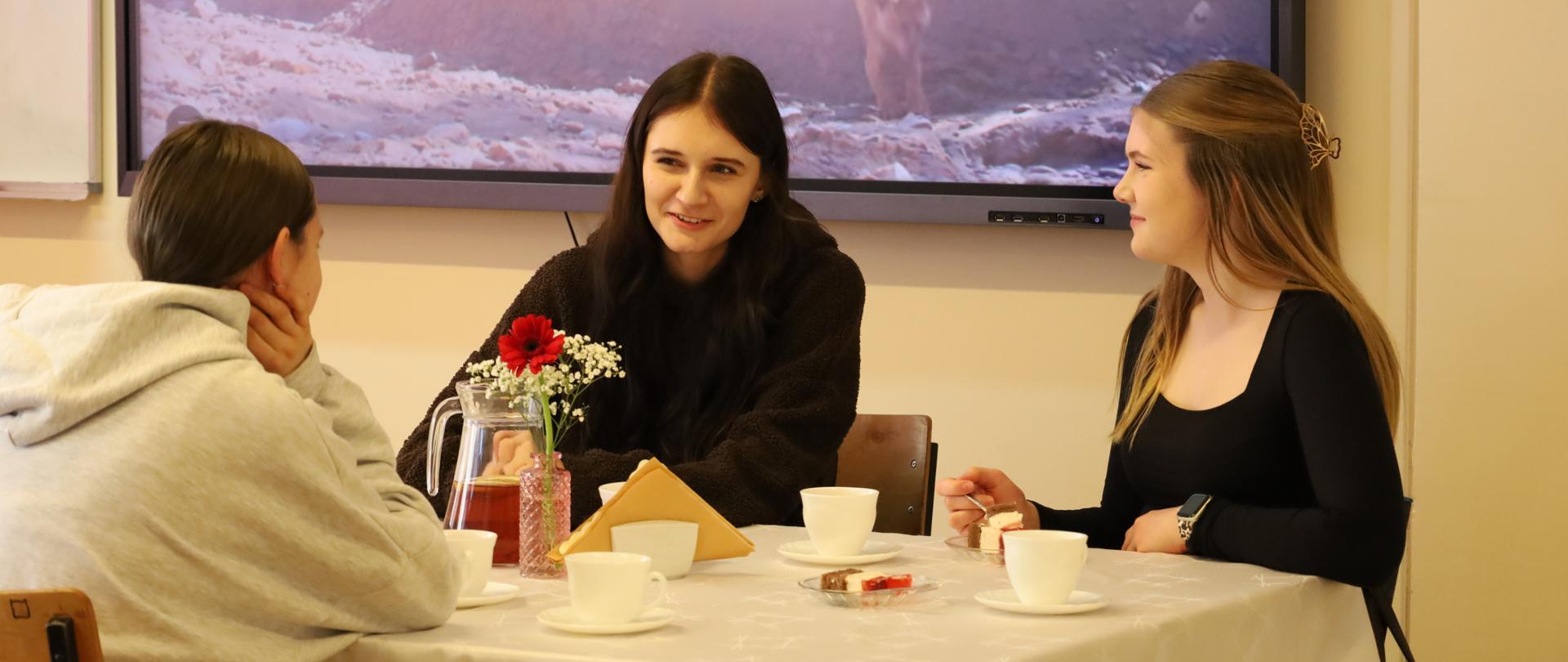 Na zdjęciu znajdują się trzy uczennice szkoły siedzące przy stoliku nakrytym białym obrusem i degustujące ciasto. W tle znajduje się monitor, na którym wyświetlany jest teledysk piosenki.