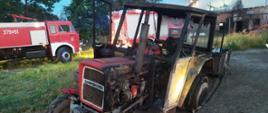 Zdjęcie przedstawia spalony ciągnik rolniczy z dwukołówką. W tle widać dwa samochody ratowniczo-gaśnicze oraz spalony budynek inwentarski