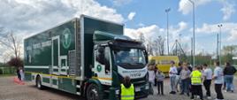 Ciężarowy pojazd Inspekcji Transportu Drogowego (IVECO) zaparkowany na placu szkolnym. Przed pojazdem stoi grupa młodzieży szkolnej oraz osoba w żółtej kamizelce odblaskowej z napisem "Kuratorium Oświaty w Opolu". W tle widoczny żółty bus Wojewódzkiego Ośrodka Ruchu Drogowego w Opolu oraz boisko sportowe.