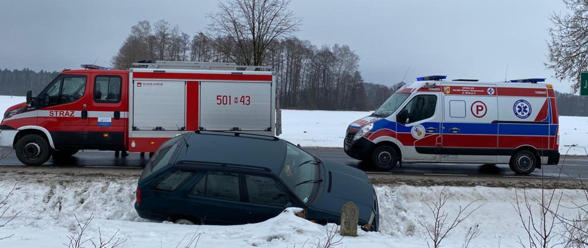 Samochód osobowy znajduje się w przydrożnym rowie. Na jednym pasie drogi stoi samochód strażacki i karetka pogotowia.