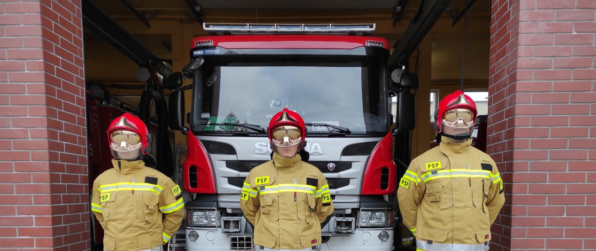 Trzech strażaków ubranych w nowe kpl. ubrań specjalnych firmy DEVA w kolorze piaskowym na tle samochodu pożarniczego