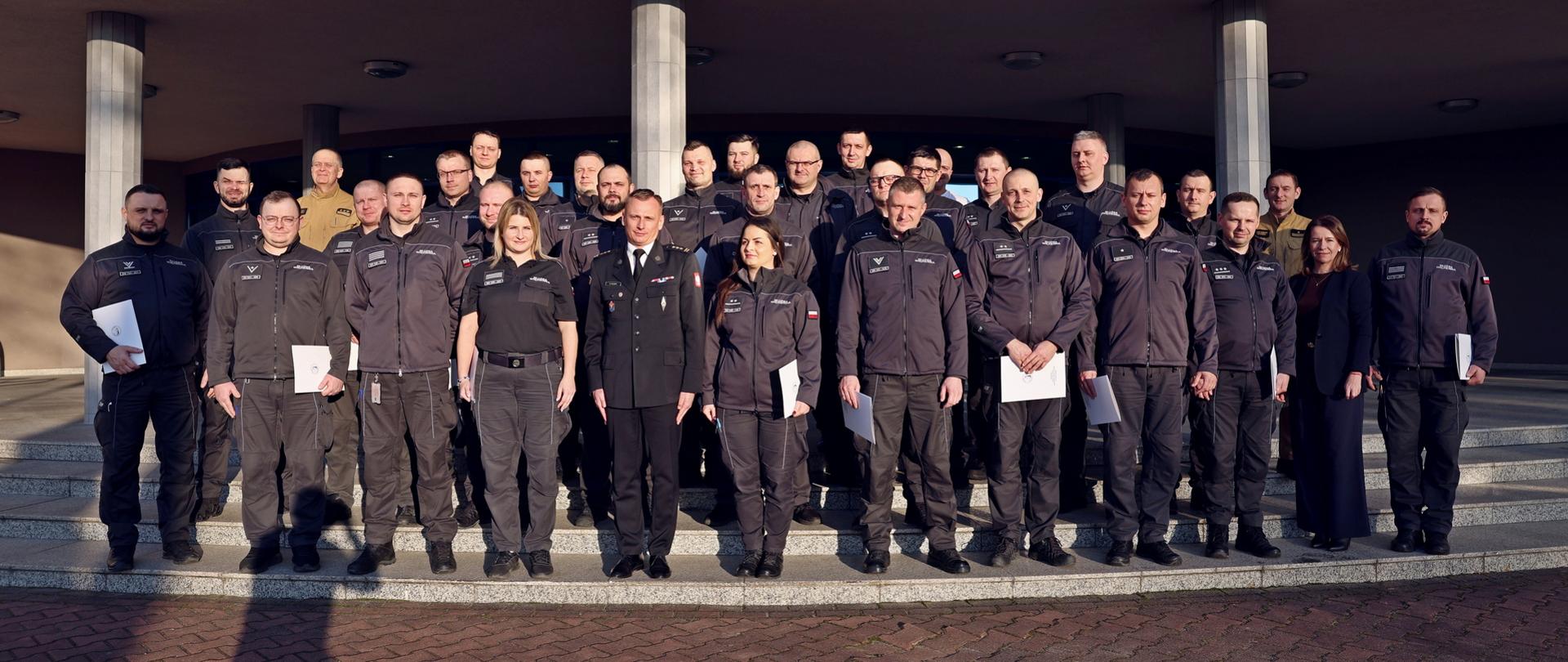 Pamiątkowe zdjęcie grupowe funkcjonariuszy i dwóch funkcjonariuszek Służby Więziennej (29 osób) będących uczestnikami szkolenia inspektorów ochrony przeciwpożarowej w towarzystwie Zastępcy Komendanta Centralnej Szkoły PSP, kobiety oraz dwóch funkcjonariuszy CS PSP
