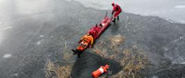 Zdjęcie przedstawia strażaków PSP ubranych w suche skafandry oraz kamizelki ratunkowe, którzy przy użyciu sań lodowych, wydobyli strażaka-pozoranta z przerębla.