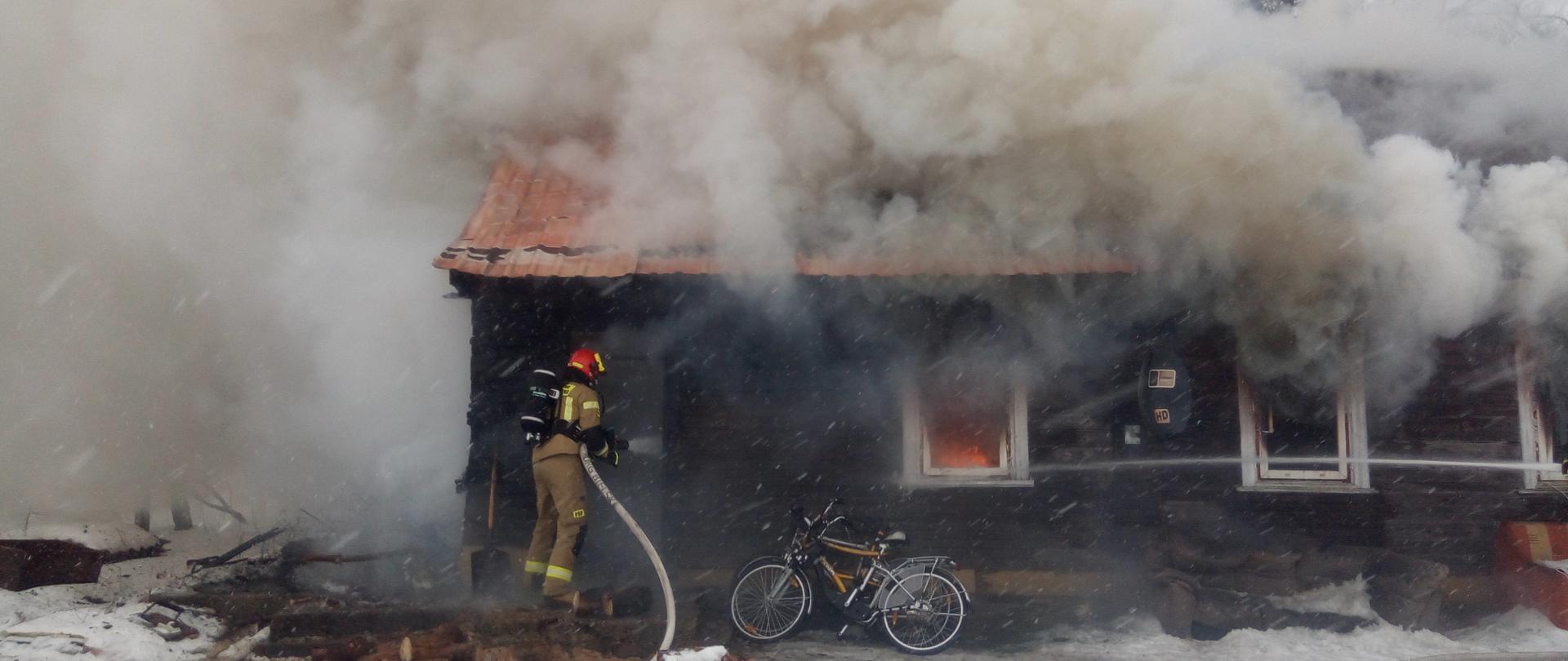 Drewniany budynek mieszkalny objęty pożarem. Przez okna wydobywa się gęsty dym, W jednym z okien widoczne płomienie. Górna część budynku zasłonięta dymem. Przed drzwiami strażak w ubraniu specjalnym koloru piaskowego, czerwonym hełmie i butlą powietrzną na plecach trzymają wąż strażacki prowadzi działania gaśnicze.