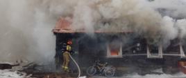 Drewniany budynek mieszkalny objęty pożarem. Przez okna wydobywa się gęsty dym, W jednym z okien widoczne płomienie. Górna część budynku zasłonięta dymem. Przed drzwiami strażak w ubraniu specjalnym koloru piaskowego, czerwonym hełmie i butlą powietrzną na plecach trzymają wąż strażacki prowadzi działania gaśnicze.