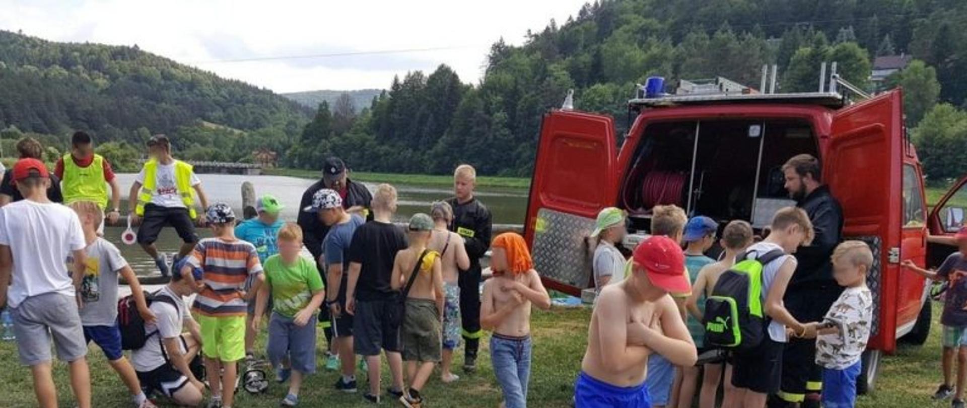 Na zdjęciu rozproszona grupa chłopców wraz ze strażakami z Ochotniczej Straży Pożarnej w tle zalew a w oddali pagórki porośnięte drzewami jeden z chłopców leży na kocu