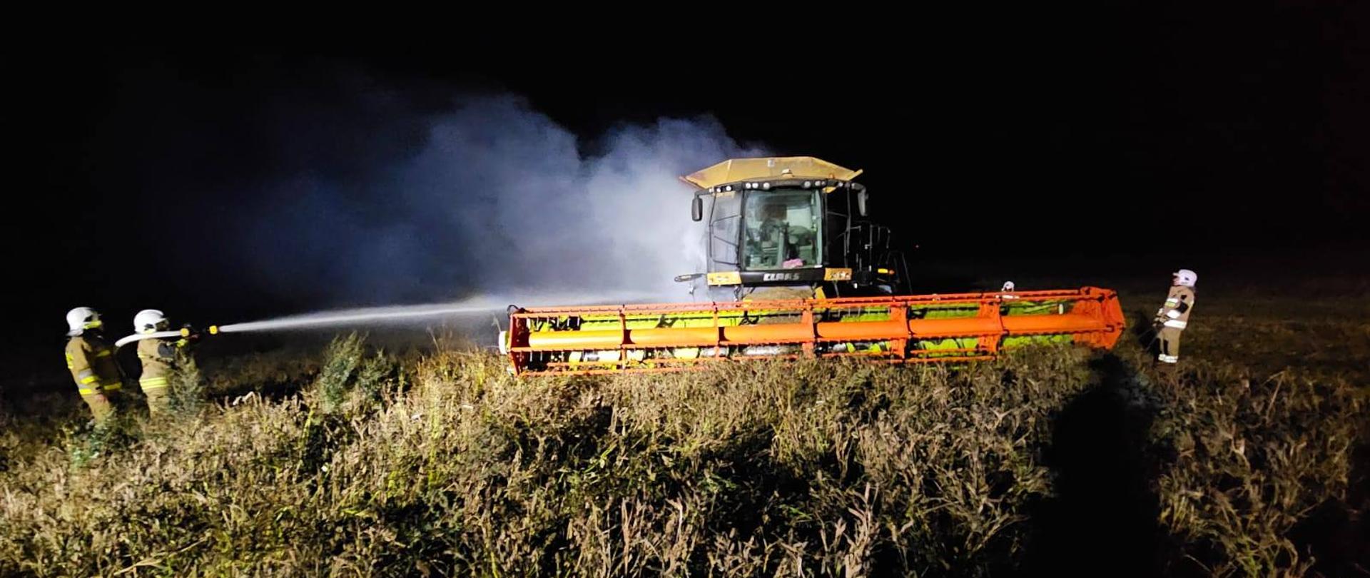 Zdjęcie zrobione nocą. Duży kombajn z czerwonym hederem stoi na polu, unosi się dym. Z lewej strony dwóch strażaków leje wodę na kombajn.