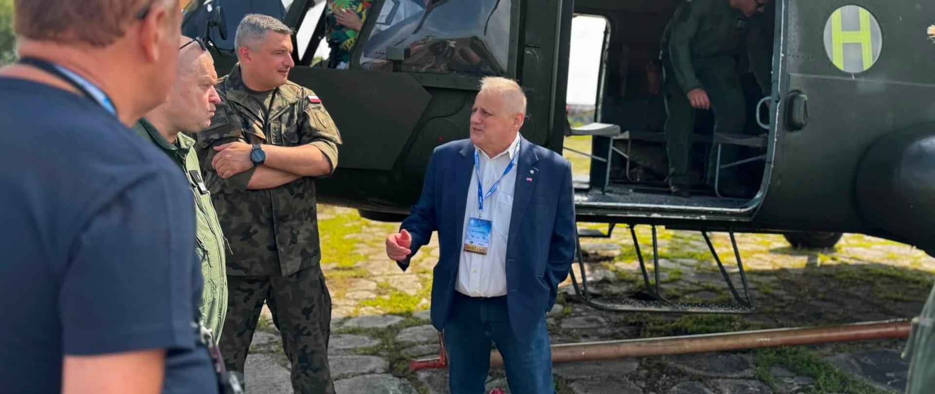 Wicewojewoda warmińsko-mazurski w garniturze rozmawia z przedstawicielami służb mundurowych na tle wojskowego helikoptera.