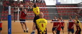 Zawodnicy grają mecz siatkówki na boisku. Dwie drużyny grają pomiędzy sobą. W tle kibice na trybunach. Zawodnicy wyskoczyli do siatki w celu przebicia piłki przez siatkę oraz obrony
