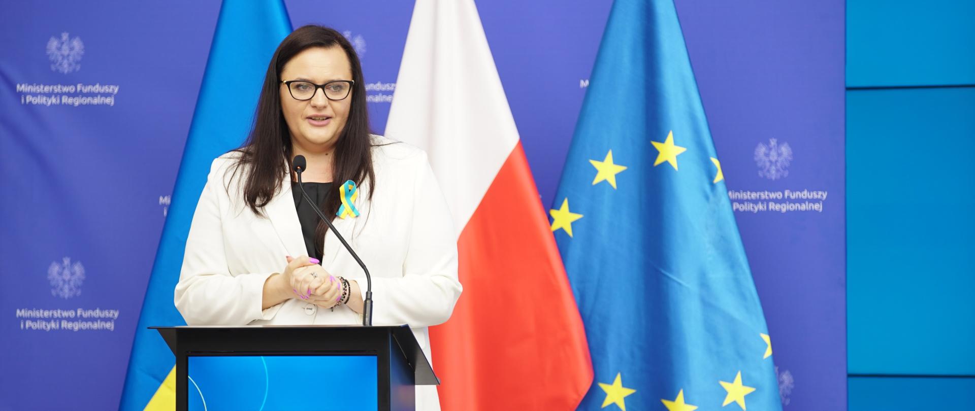 Wiceminister Małgorzata Jarosińska-Jedynak w sali konferencyjnej w mównicy. Za nią flago UA, PL i UE.
