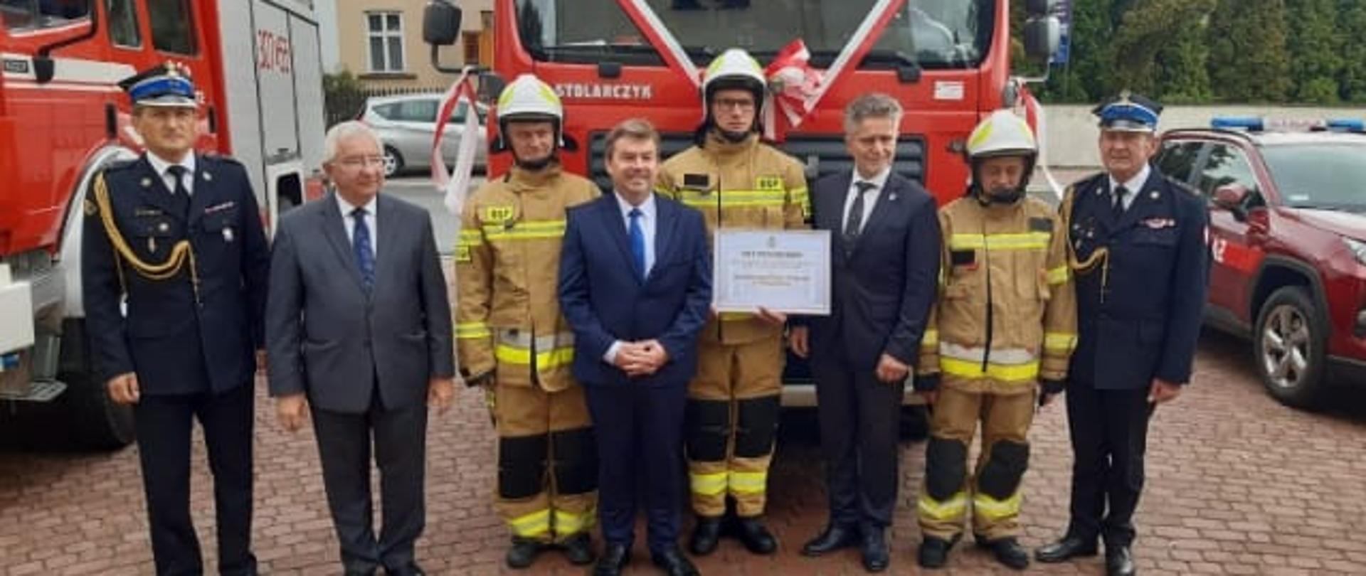 Zdjęcie przedstawia zaproszonych gości oraz druhów stojących na tle pozyskanego ciężkiego samochodu gaśniczego.