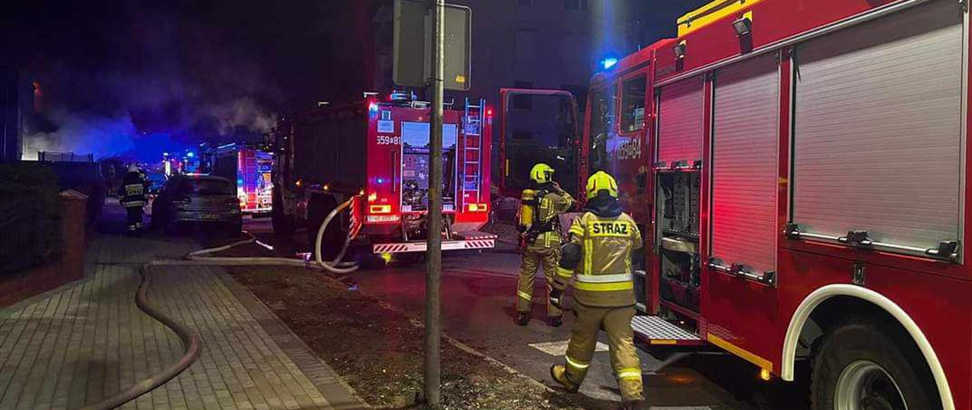 Zdjęcie przedstawia dwa samochody pożarnicze stojące na ulicy. Przy jednym samochodzie stoi dwóch strażaków, na chodniku rozciągnięte są węże pożarnicze.