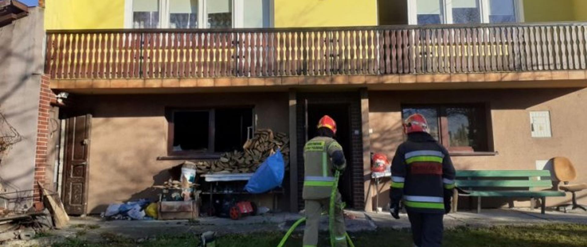 Zdjęcie przedstawia dwóch strażaków w ubraniach specjalnych i hełmach wchodzących do domu z linią gaśniczą. Wejście na parterze, po lewej stronie widać otworzone okna oraz ułożone na zewnątrz różne przedmioty gospodarstwa domowego. Budynek wielokondygnacyjny.