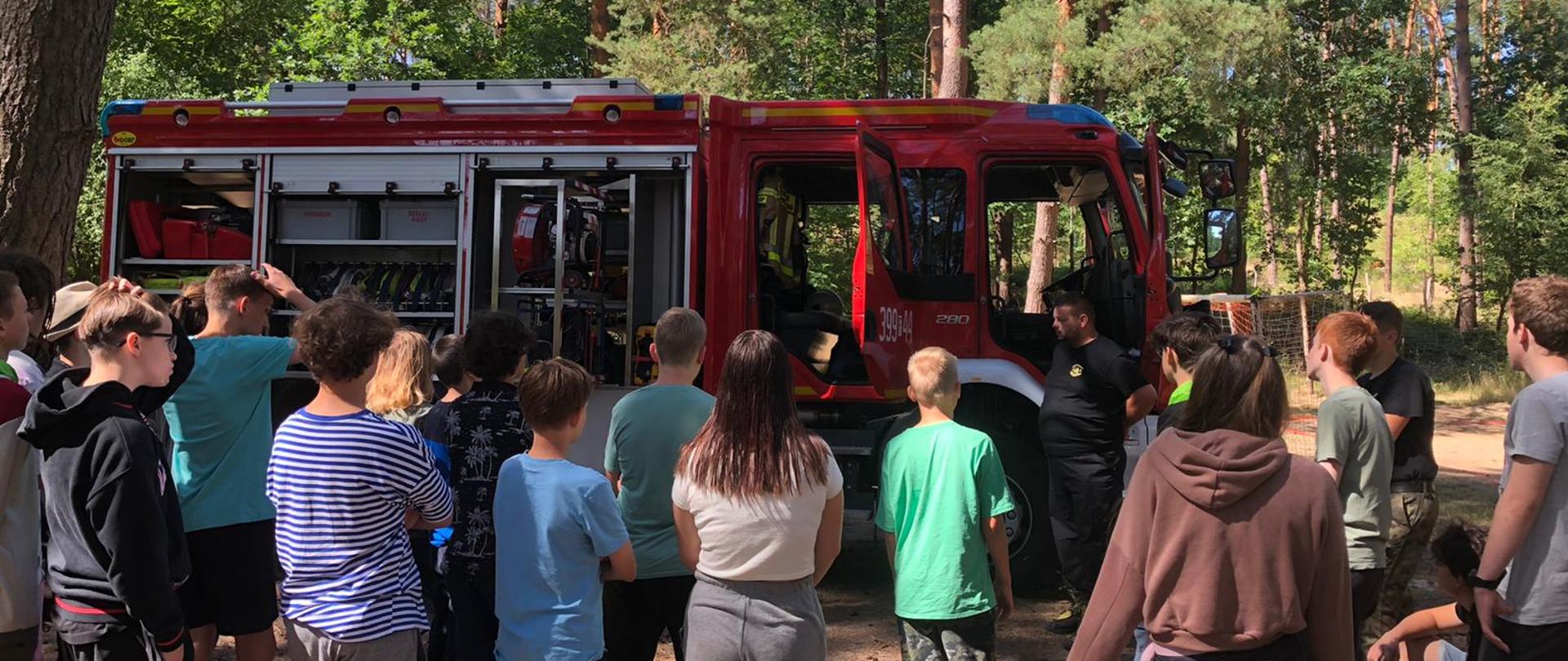 Grupa stojącej młodzieży na tle samochodu pożarniczego w lesie. Przed samochodem pożarniczym umundurowany strażak prezentujący sprzęt na samochodzie.