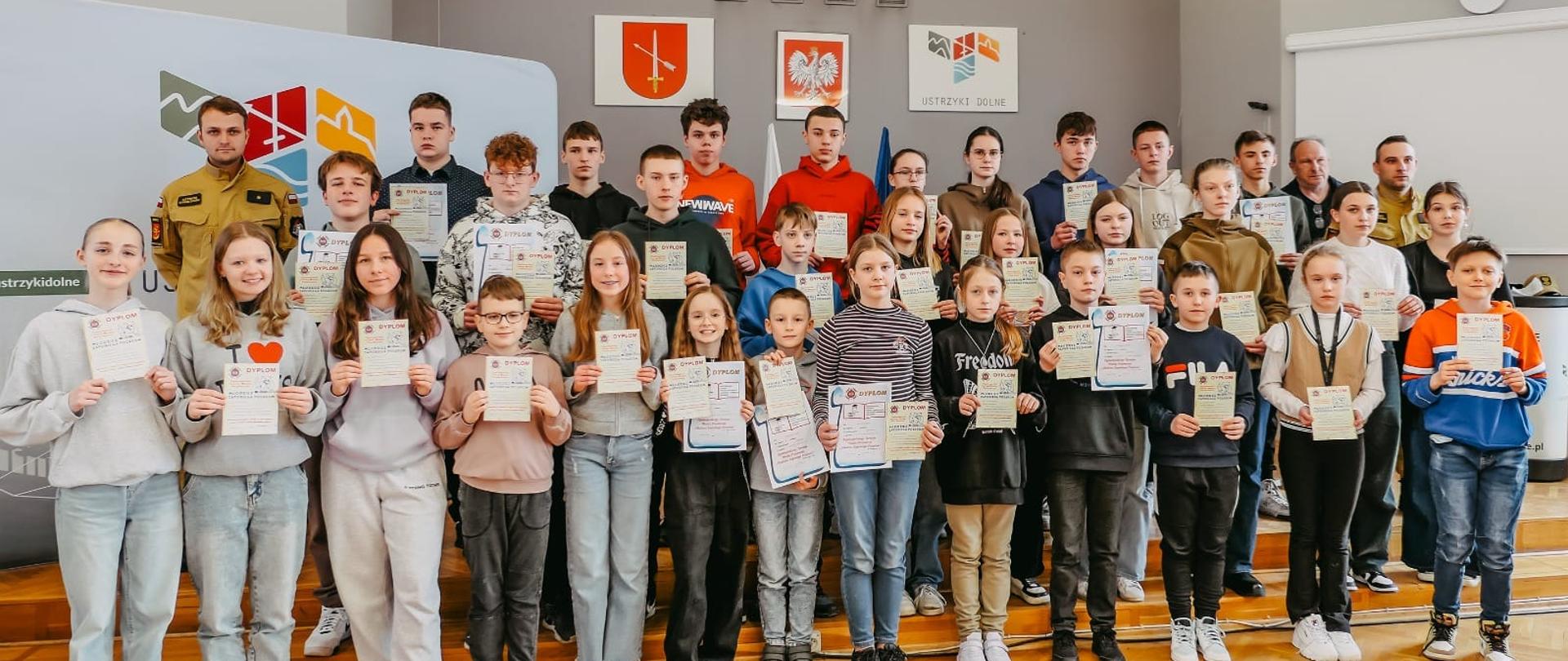 Zdjęcie przedstawia dwóch funkcjonariuszy psp i członków komisji konkursu a także wszystkich uczniów którzy brali udział w konkursie z dyplomami i nagrodami w rękach. 