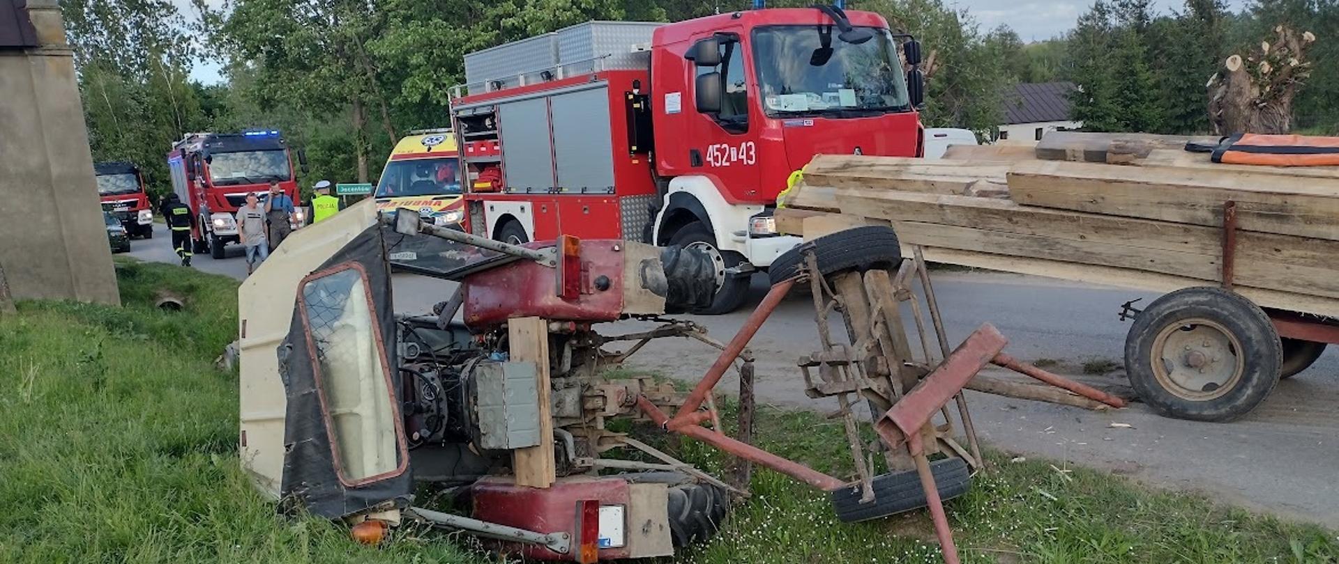 Ciągnik z kabiną leży wywrócony w przydrożnym rowie na prawym boku. Na jezdni stoi wóz z drzewem oraz samochód gaśniczy i karetka pogotowia. W tle strażak, policjant i osoby postronne. 