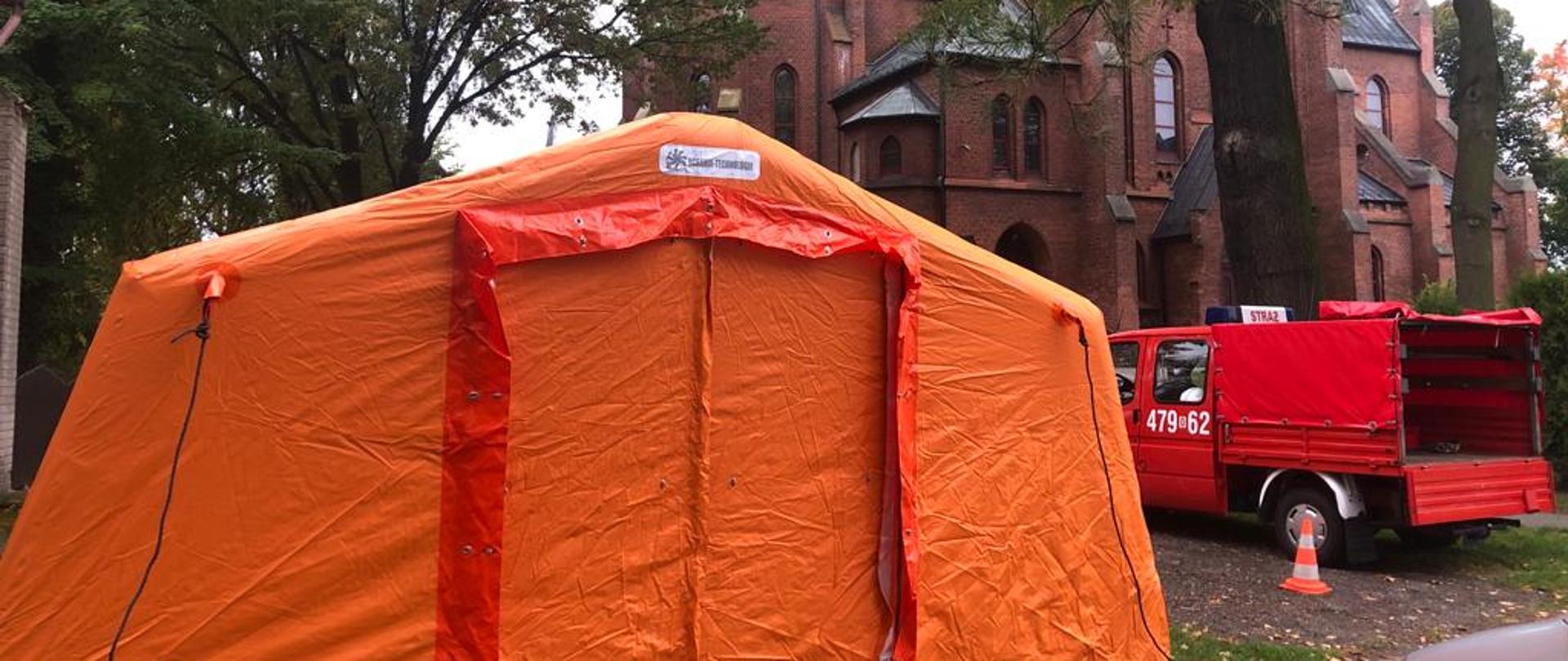 Na zdjęciu namiot koloru pomarańczowego po prawej stronie samochód pożarniczy w tle Kościół.
