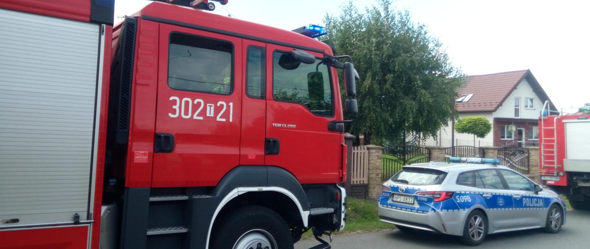 Zdjęcie przedstawia zaparkowane na drodze samochody służb ratowniczych. Kolejno stoją : samochód gaśniczy, radiowóz i drugi samochód strażaków. 