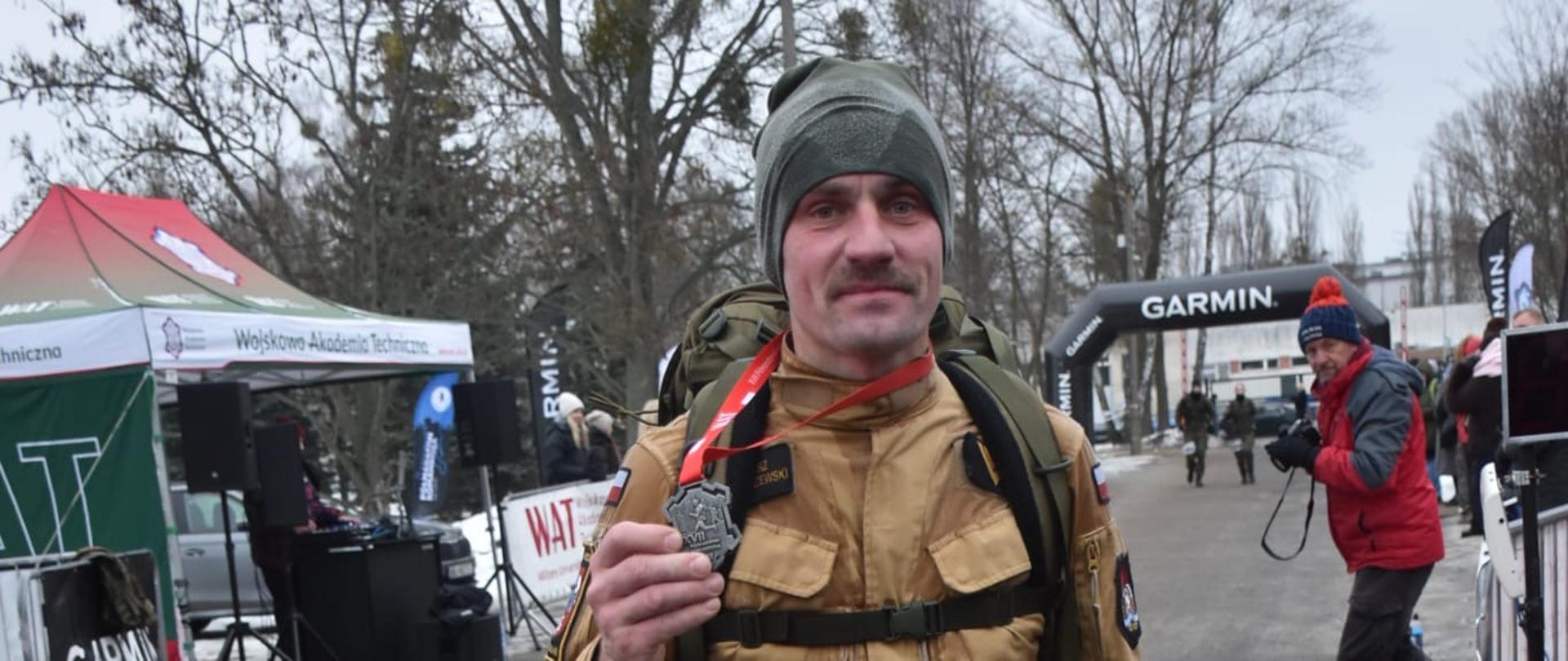 Na pierwszym zdjęciu widać mężczyznę stojącego na mecie biegu plenerowego. Ma na sobie beżowy mundur, czapkę typu beanie oraz duży plecak z pasami piersiowymi i biodrowymi. Na szyi zawieszony jest medal, który trzyma w dłoni i pokazuje do zdjęcia. Na udzie ma przypięty numer startowy 361. W tle znajdują się namioty organizatorów, brama z logo Garmin oraz inne osoby uczestniczące w wydarzeniu. Pogoda jest chłodna, drzewa są bez liści.