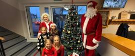 Na szkolnym korytarzu Święty Mikołaj w czerwonym stroju obok niego grupa uczniów oraz dwie kobiety blondynku w okularach ubrane na czerwono. W tle widać świąteczną choinkę oraz szkolne wyposażenie.