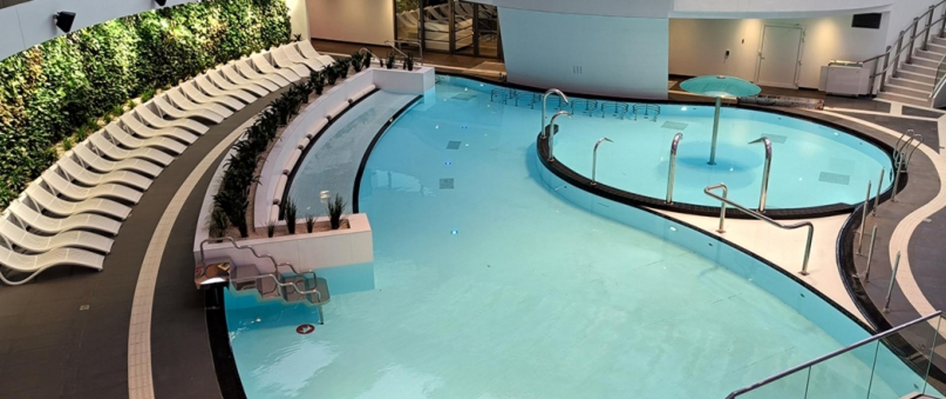 Wnętrze basenu z leżakami, zieloną ścianą i wodnym parasolem; nowoczesny wystrój i przeszklone elementy.
