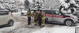 Kolorowe zdjęcie przedstawia na środku, bok srebrnego samochodu pożarniczego Państwowej Straży Pożarnej z napisem Straż na czerwonym pasem. Obok pojazdu czterej umundurowani strażacy Państwowej Straży Pożarnej i Ochotniczej Straży Pożarnej pomagają starszej osobie wyjść z pojazdu. Na całym planie fotografii widoczne mocno zaśnieżone pojazdy osobowe na parkingu i duża ilość śniegu. W tle zaśnieżone drzewa.