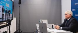 Wiceminister Maciej Lasek podczas łączenia online na konferencji prasowej, minister siedzi przy stole z laptopem, przed nim ekran z uczestnikami konferencji w Katowicach 