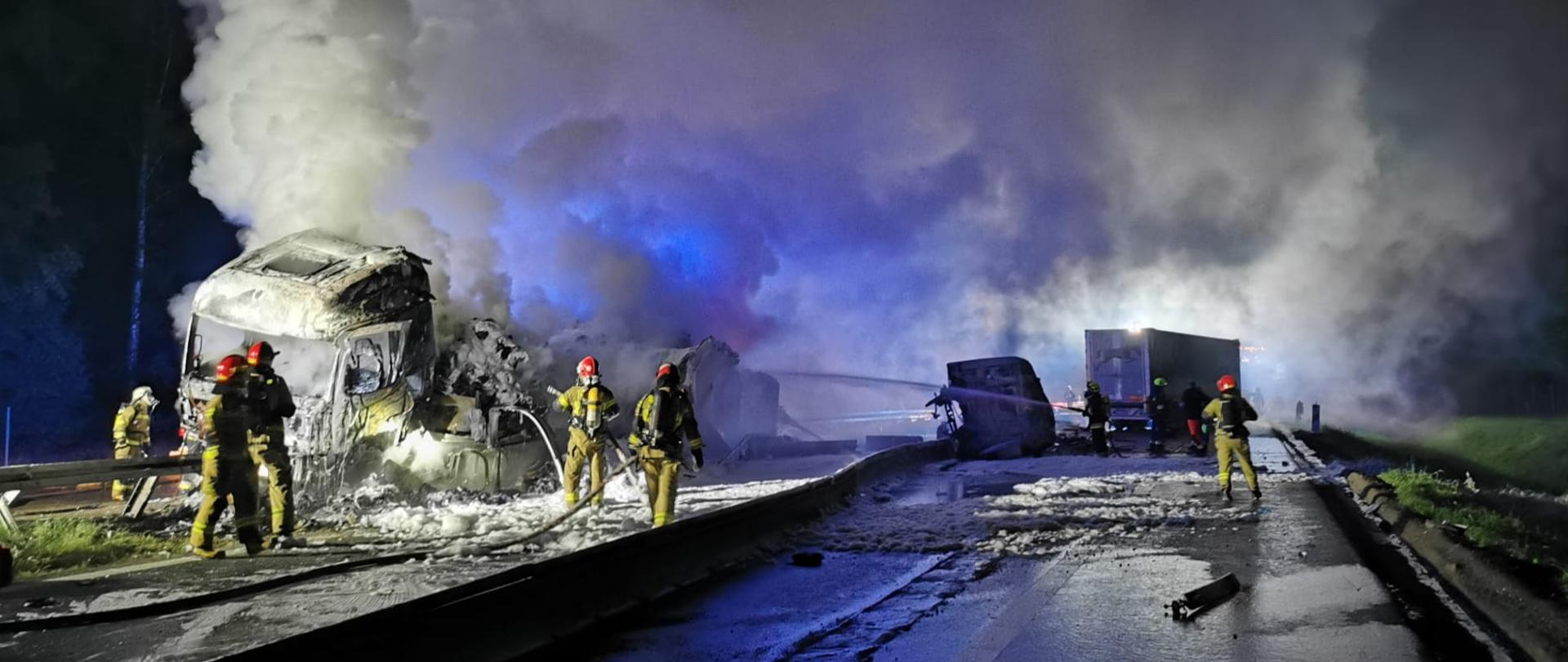 Na zdjęciu znajdują się wraki spalonych samochodów ciężarowych na autostradzie, nad którymi unoszą się kłęby pary i dymu oraz strażacy podający pianę gaśniczą 