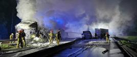 Na zdjęciu znajdują się wraki spalonych samochodów ciężarowych na autostradzie, nad którymi unoszą się kłęby pary i dymu oraz strażacy podający pianę gaśniczą