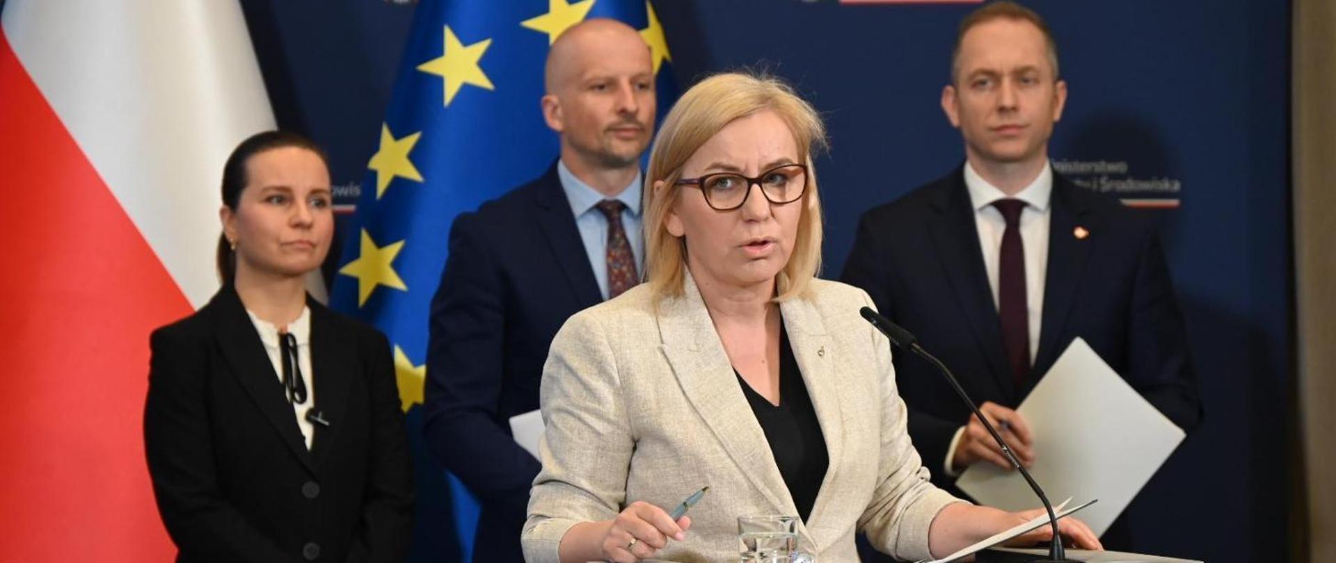 Minister of Climate and Environment Paulina Hennig-Kloska during a joint press conference on the environmental component of the East Shield, together with Deputy Minister of National Defence Cezary Tomczyk, Deputy Minister of Climate and Environment Urszula Zielińska, and Chief Nature Conservator Mikołaj Dorożała. In the foreground, Minister Paulina Hennig-Kloska is seen speaking into a microphone. Behind her stand the other participants of the conference. In the background on the right, the Polish flag and the European Union flag are visible.