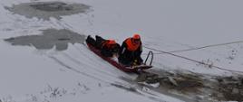 Przerębel. Dwóch strażaków ubranych w suche skafandry znajdują się na saniach lodowych są siciagani za pomocą liny w stronę brzegu stawu.
