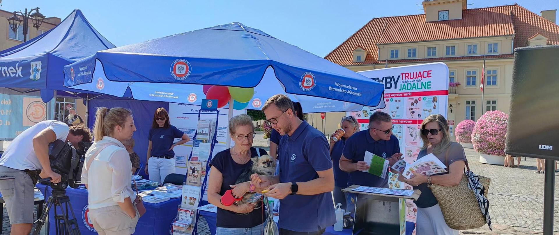Stoisko profilaktyczne podczas akcji "Zdrowe wakacje". Niebieski namiot, stół z ulotkami, edukatorzy i zainteresowane osoby.