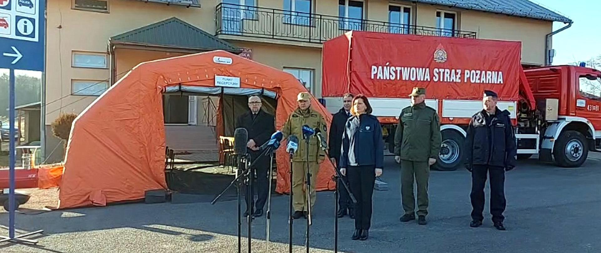 Zdjęcie zrobione na zewnątrz. Na pierwszym planie sześcioro uczestników konferencji prasowej. Wśród nich oficerowie: Państwowej Straży Pożarnej, Policji i Straży granicznej ubrani w mundury. w tle namiot strażacki z tablicą informacyjną „punkt recepcyjny”. bok stoi ciężarowy samochód, na którym jest napis Państwowa Straż Pożarna.