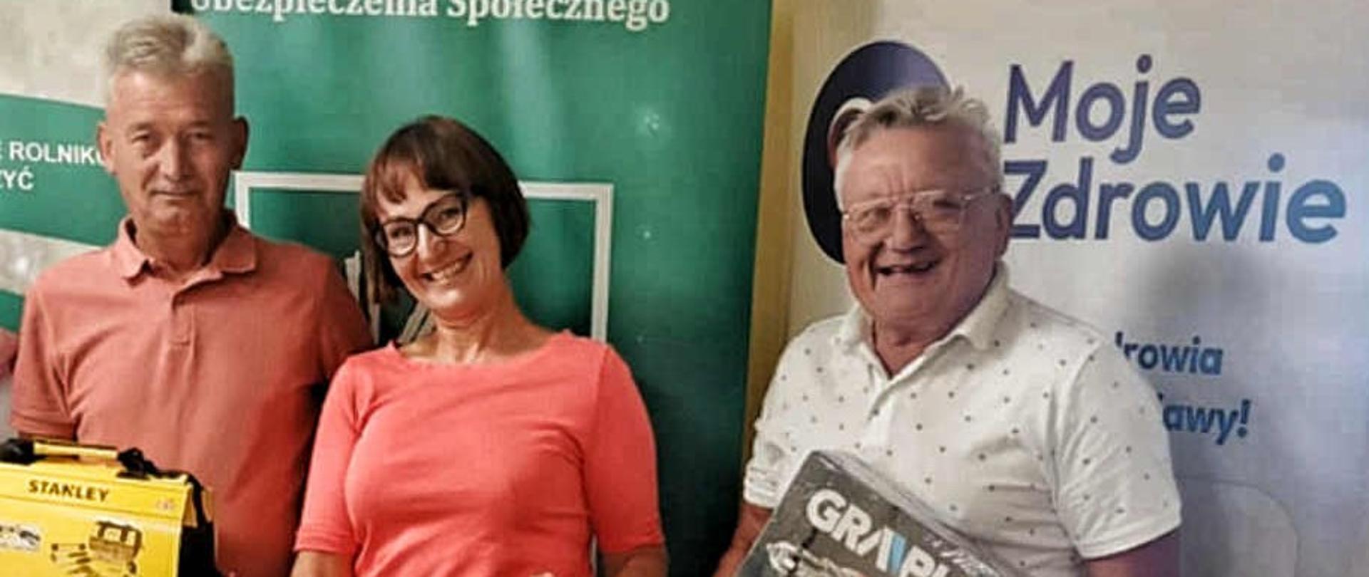 Trzy osoby, kobieta i dwóch, mężczyzn pozują do zdjęcia na tle rollupu KRUS, uśmiechają się, pokazując swoje nagrody – skrzynki z narzędziami.