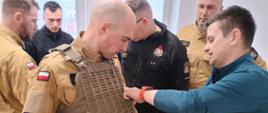 Mężczyzna zakłada kamizelkę balistyczną strażakowi w mundurze koloru musztardowego obok stoją inni strażacy w mundurach służbowych koloru musztardowego oraz czarnego.
