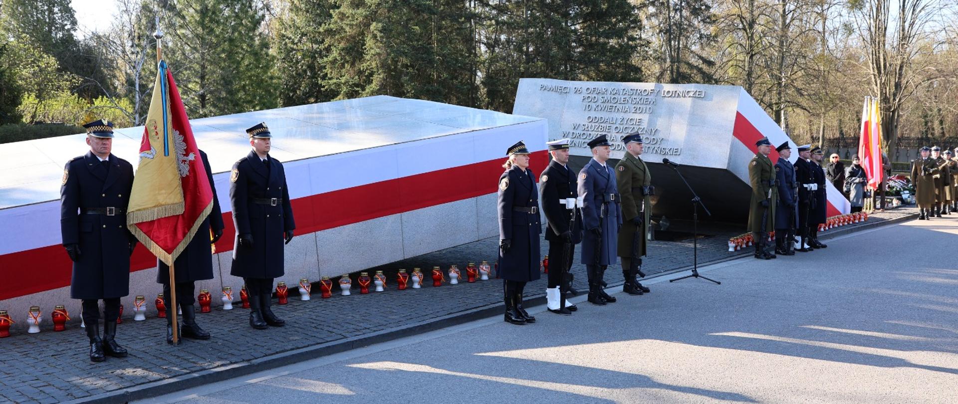 Uroczystość przy pomniku upamiętniającym ofiary katastrofy smoleńskiej – funkcjonariusze służb mundurowych stoją w szeregu przy biało-czerwonym memoriale, obok ustawiono znicze. W tle widoczny jest las, a przed pomnikiem przygotowano miejsce do składania wieńców.